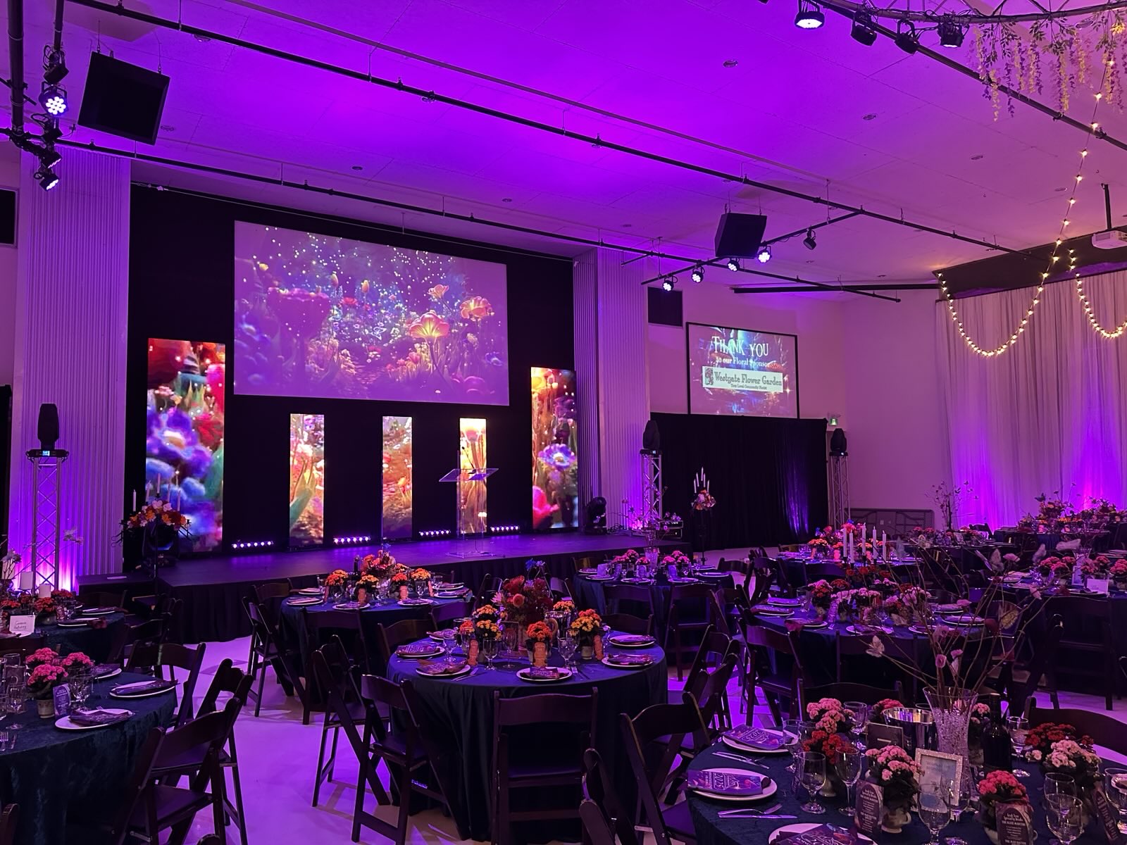 Gala dinner room washed in purple uplighting with floral centrepieces, banquet tables and a large stage screen — event lighting and staging by Valley Tech
